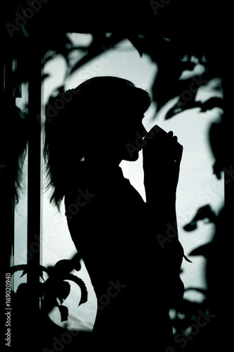 silhouette of the figure of a woman drinking hot tea near a window in foliage of a tree