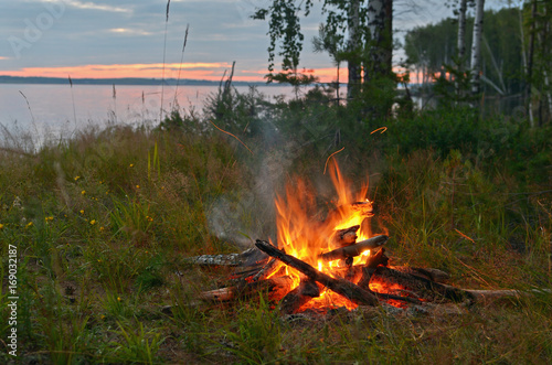 Fototapeta Naklejka Na Ścianę i Meble -  Summer evening campfire by the river against the setting sun.