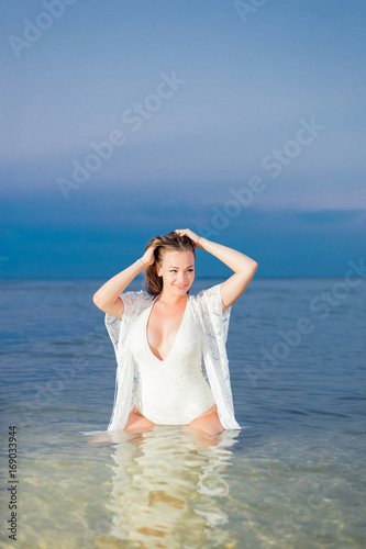 Foto Beautiful sexy girl in a white bathing suit and tunic sits in the water near the shore