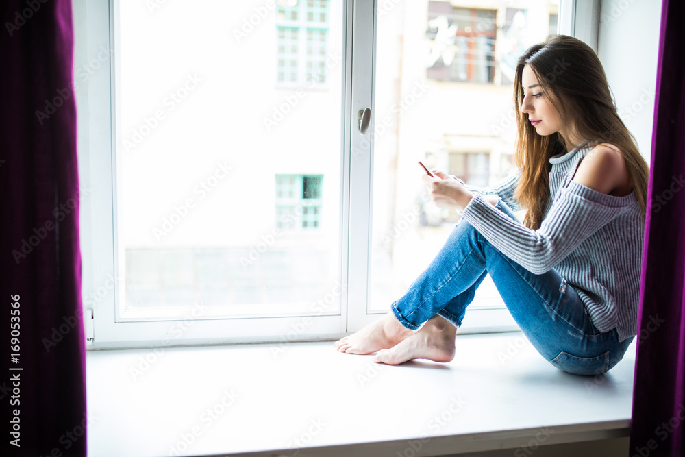 young woman with cellphone in hands siting on window sill young woman with cellphone in hands siting on window sill