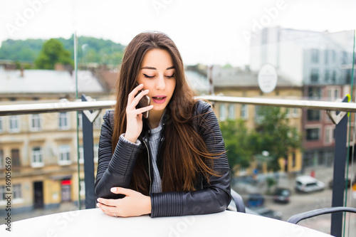 Young cute lady is talking on her smart phone while having coffee in the open air terrace of cafe.