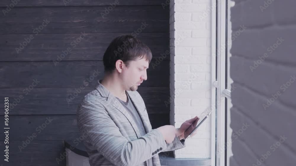 Young man working on tablet. Confident young man working on digital tablet while standing in front of the big window in modern office Young man working on tablet. Confident young man working on digital tablet while standing in front of the big window in modern office