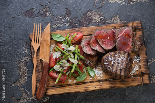 Grilled medallion beefsteaks with asparagus and tomato salad on a wooden serving tray. Flat-lay on a brown stone background, horizontal shot