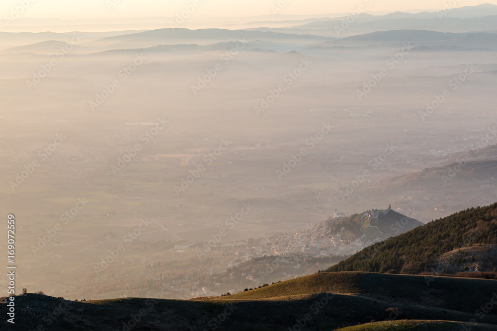 An aerial view of Assisi town and St.Francis church over a sea of mist at sunset, with warm and soft colors