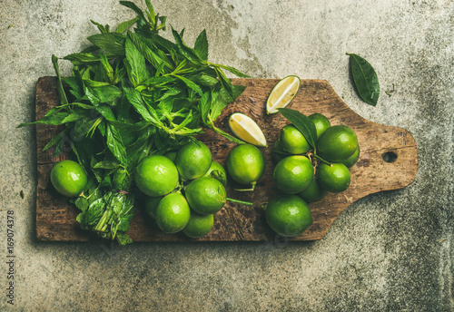 Flatlay of freshly picked organic limes and mint leaves for making cocktail or lemonade on wooden rustic board over grey concrete stone background, top view