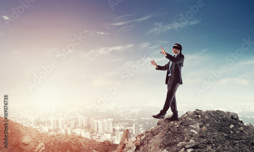 Young businessman in blindfold walking carefully and cityscape at background