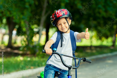 Little boy learns to ride a bike in thepark near the home. Kid shows the thumbs up on bicycle. Happy smiling child in helmet riding a cycling.
