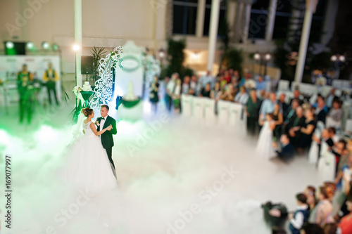 Beautiful wedding couple dancing their first dance in the huge hall with heavy smoke, different lights and people looking at them.