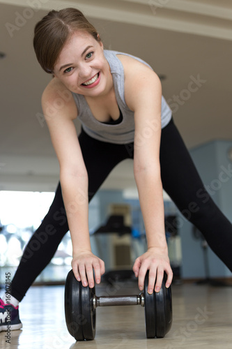 Portrait of smiling pretty girl lifting heavy weight dumbbell in front of her looking happy, strong and sharp suggesting healthy and fit lifestyle with training apparatus and gym on background