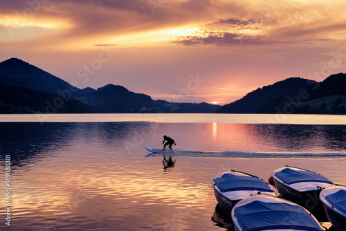 Sunset at Lake Fuschl with silhouette of a man riding an electric surfboard, boats in foreground, Fuschl am See, Fuschlsee, Salzburg state, Salzkammergut, Austria, Europe © dinkaspell