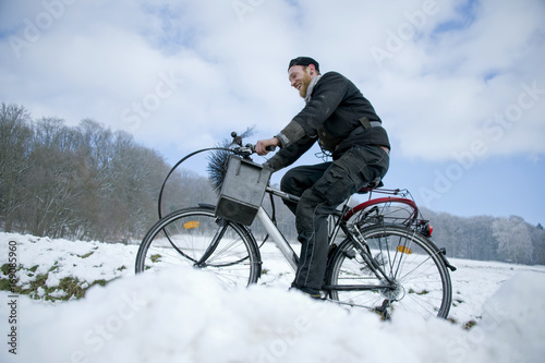 Schornsteinfeger fährt mit Fahrrad und Arbeitsgerät durch verschneite Winter Landschaft
