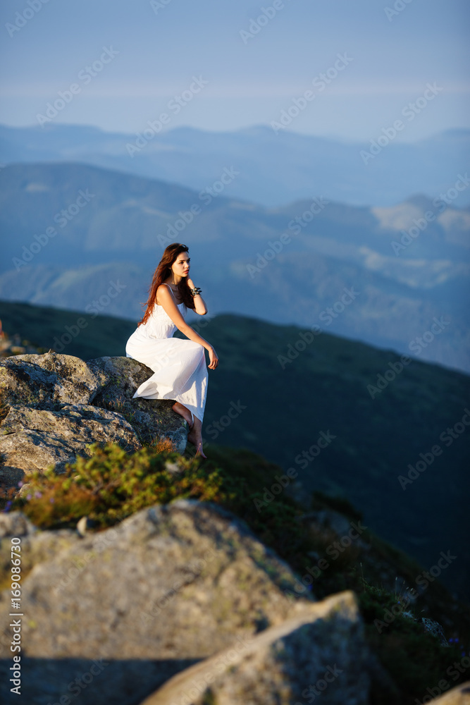 Naklejka premium Beautiful girl with slender figure in a long dress profile. Portrait of a young beautiful woman on the nature. Pretty young woman sits on a rock. Beautiful girl posing in high mountain scenery