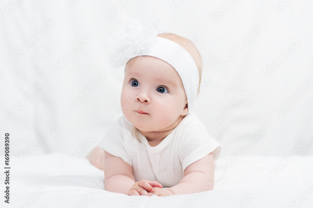 Cute baby girl in white shirt, with bow flower on head, lying on belly on soft blanket, indoors Cute baby girl in white shirt, with bow flower on head, lying on belly on soft blanket, indoors