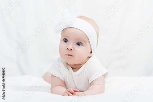 Cute baby girl in white shirt, with bow flower on head, lying on belly on soft blanket, indoors