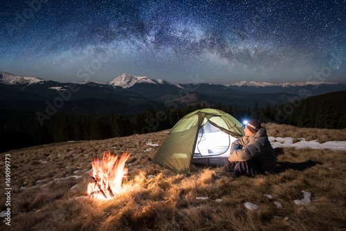Male tourist enjoying in his camp at night. Man with a headlamp sitting near campfire and tent under beautiful sky full of stars and milky way. On the background snow-covered mountains