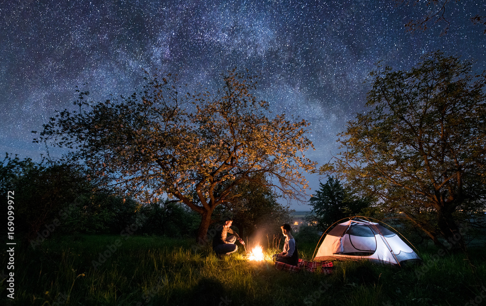 Plakat Romantic couple tourists sitting at a campfire near tent under trees and beautiful night sky full of stars and milky way. Night camping