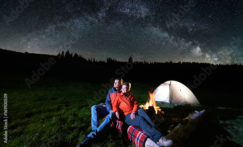 Night camping in the mountains. Romantic couple tourists sitting at a campfire near illuminated tent under beautiful night sky full of stars and milky way. Long exposure