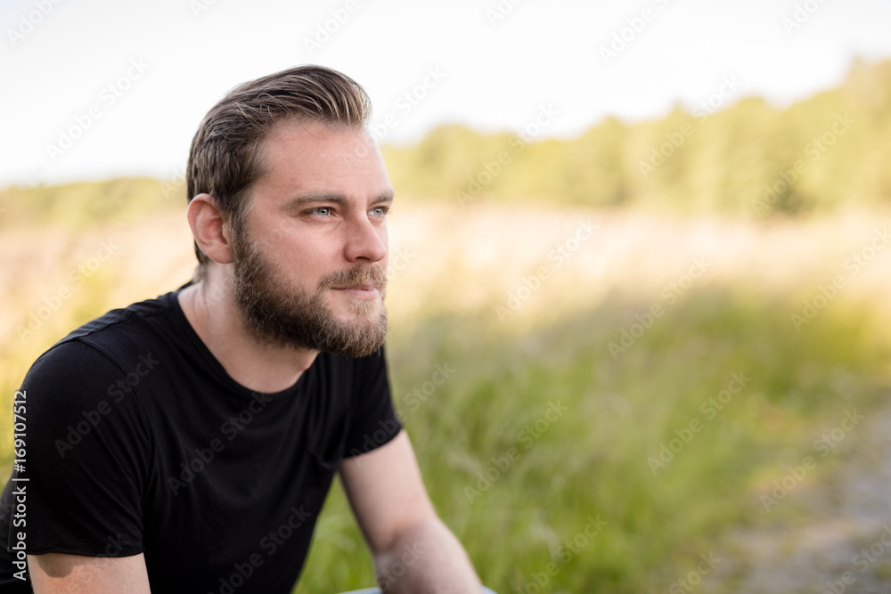 Laid back bearded man wearing a black shirt, standing outdoors on a sunny summer day looking away from camera with a smile on his face.