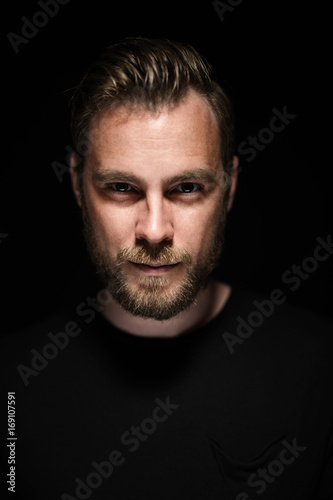 Portrait of a bearded man with an intense look standing in a dark room against a black background.