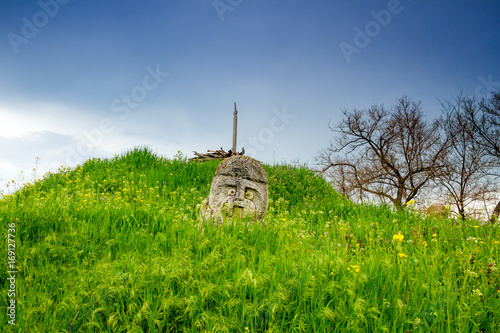 Scythian warrior stone statue and wooden sword on top of curgan with fresh green grass and blue cloudy sky in background, Khortytsia island, Zaporozhye, Ukraine photo