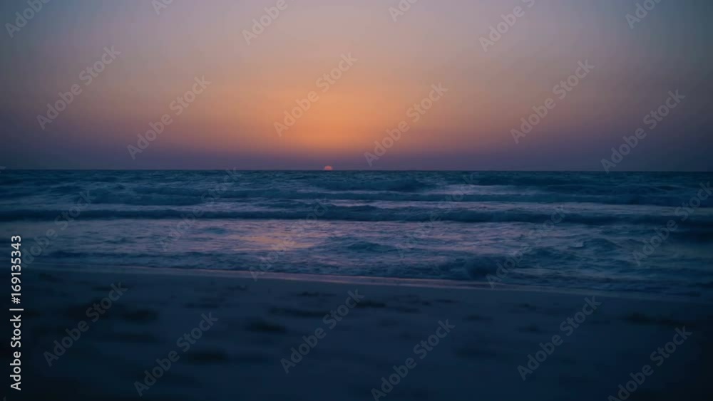 Amazing mystical bloody sunset on the sea. Clear sky. The calm sea with small waves. Waves rippling over sand. View from the beach to horizon. Sun sets over the sea. Half of sun is submerged in sea Amazing mystical bloody sunset on the sea. Clear sky. The calm sea with small waves. Waves rippling over sand. View from the beach to horizon. Sun sets over the sea. Half of sun is submerged in sea