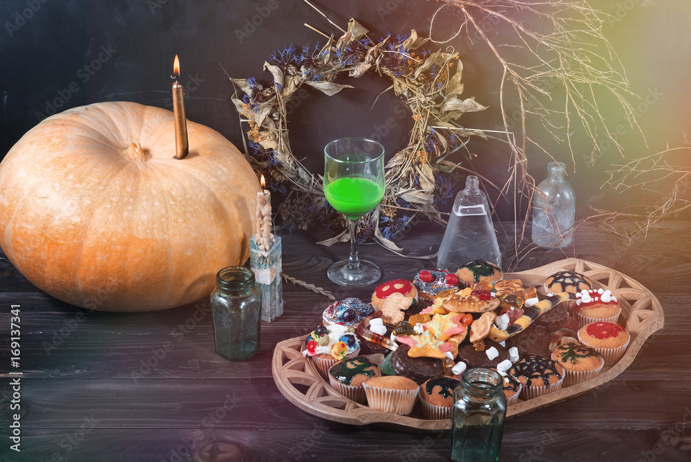 Pumpkin, candle, green liquid in a glass, sweets, cookies, jars, dry twigs and leaves on a table