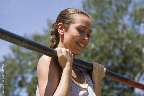 Young  brunette woman doing pull-up on a sports horizontal bar at summer day photo