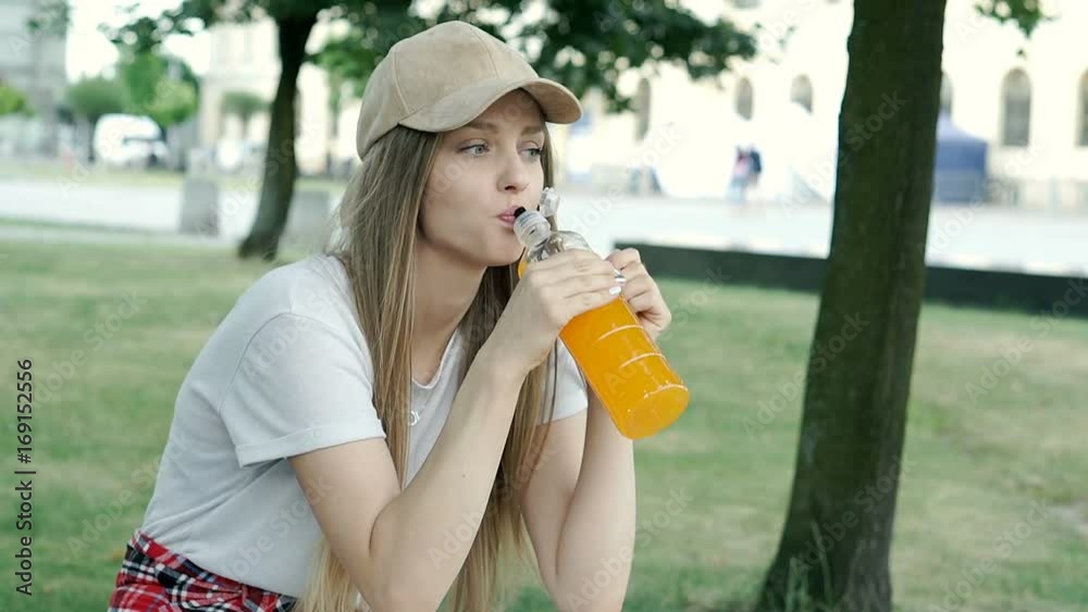 Pretty girl in cap drinking beverage in the park and smiling to the camera, steadycam shot

