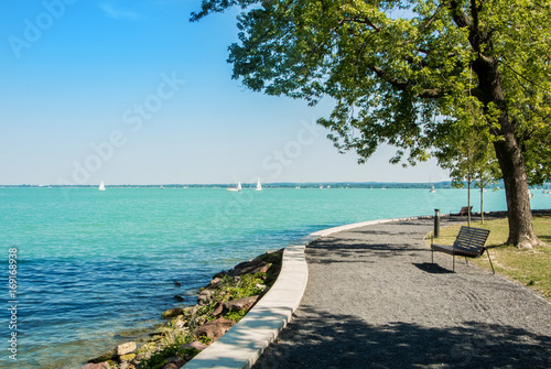 A view of Balaton lake with white yachts at the horizon and trees, bench and footpath at the foreground, Tihany, Hungary.