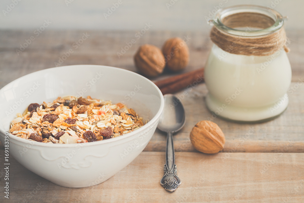 White bowl with Granola or Muesli and a jar of milk or plain yogurt and a spoon on a vintage wood background. Healthy breakfast composition. White bowl with Granola or Muesli and a jar of milk or plain yogurt and a spoon on a vintage wood background. Healthy breakfast composition.