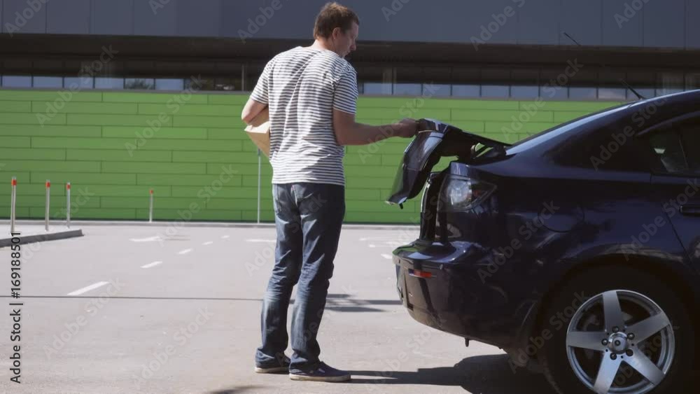 Handsome guy after visiting grocery store. Happy casual guy putting purchases into trunk car on parking near shopping mall. Male wearing in casual clothes in sunny day outdoors. Handsome guy after visiting grocery store. Happy casual guy putting purchases into trunk car on parking near shopping mall. Male wearing in casual clothes in sunny day outdoors.