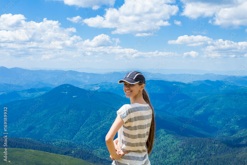 young tourist girl with long hair in a cap on top of the mountains, beautiful mountains and clouds on the background