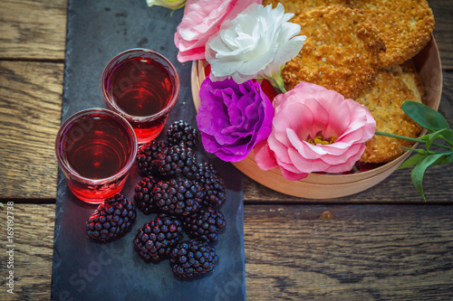 Fototapeta Naklejka Na Ścianę i Meble -  Homemade berry liqueur with fresh ripe blackberries, colorful eustoma flowers and sesame biscuits  in a wooden box and black stone tray on wooden background. Sweet alcohol drink.