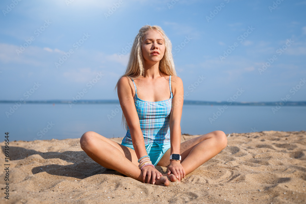 Blonde long haired girl in a blue suit is stretching and doing yoga on a lovely beach in sunlight of the rising sun Blonde long haired girl in a blue suit is stretching and doing yoga on a lovely beach in sunlight of the rising sun