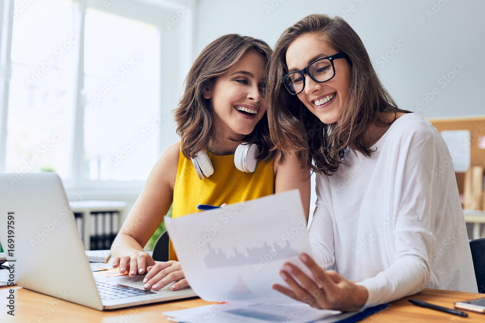Couple of cheerful ladies working laughing while working together on laptop and documents in office Couple of cheerful ladies working laughing while working together on laptop and documents in office