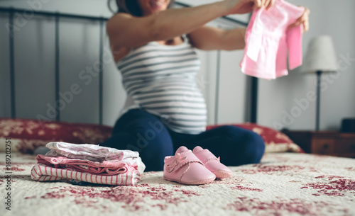 Unrecognizable pregnant woman looking baby girl cardigan. Selective foreground focus on baby clothes and shoes photo