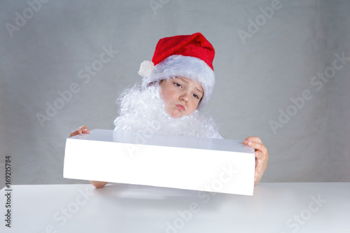 Little Santa returns a gift in a white box. Sits by the table. Close-up. Gray background. © kravik93