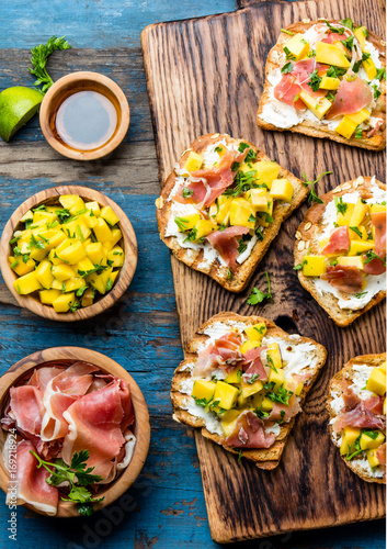 Toasts with cream cheese, ham jamon serrano and mango served on wooden board with red wine, blue wooden rustic background, top view