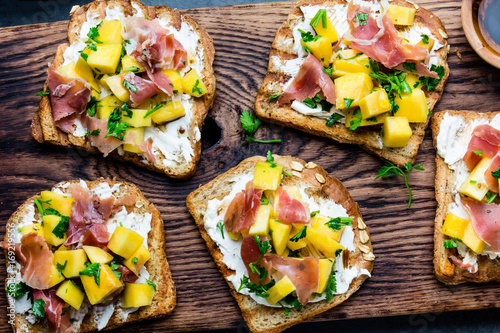 Toasts with cream cheese, ham jamon serrano and mango served on wooden board with red wine, gray slate background, top view