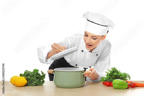 Young female chef near table with saucepan and different products, on white background