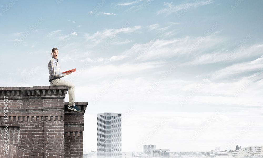 Man on roof edge reading book and cityscape at background Man on roof edge reading book and cityscape at background