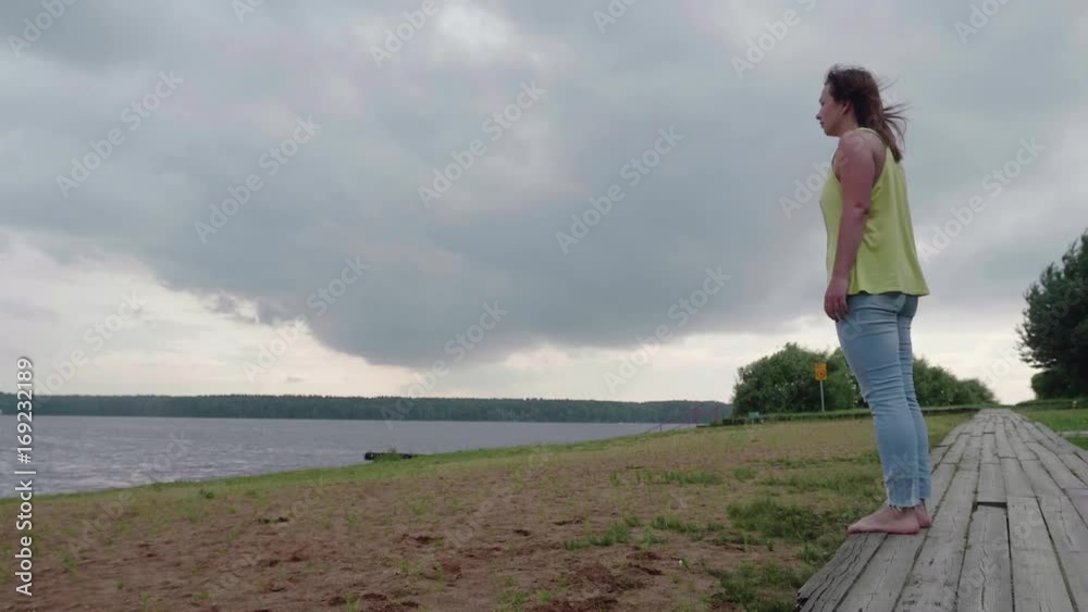 Slow motion of girl in yellow tank and blue jeans leaps for joy on bank of river on cloudy summer day Slow motion of girl in yellow tank and blue jeans leaps for joy on bank of river on cloudy summer day