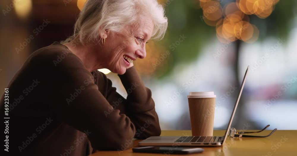 Slowmo of cheerful mature woman sitting outside coffee shop watching funny video on laptop. Lovely senior woman in outdoor setting watching video on computer smiling and laughing. 4k 