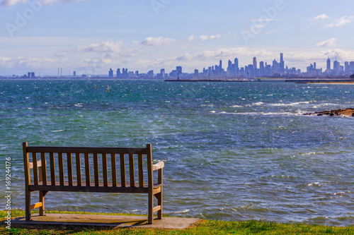 Empty wooden bench on ocean shore overlooking the skyline of Melbourne CBD on bright sunny day