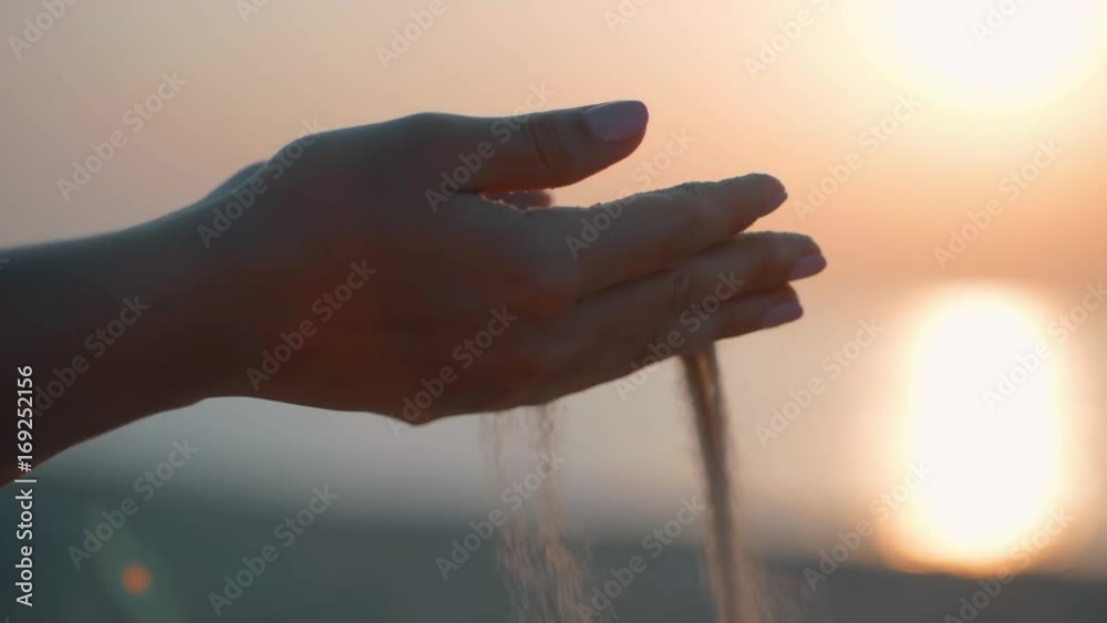 Close up of woman pouring sand running through fingers slow motion at the beach with sun flare and blue sky. Beautiful scene of young lady playing with sand on sunrise or sunset.