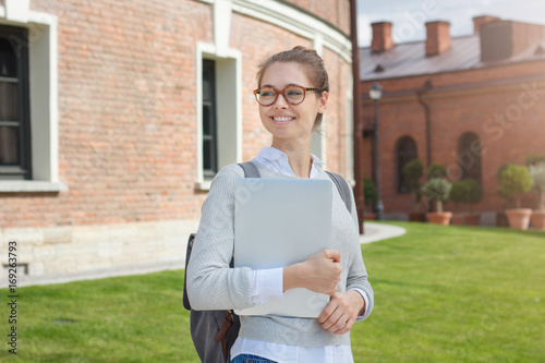Outdoor portrait of young good-looking female studying in university standing in yard with backpack and tablet PC in hands, looking leftwards as if waiting for someone to attend lectures together. photo