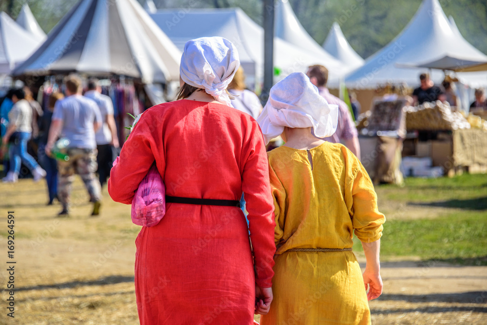 Backs of two young women in the medieval dresses and white kerchiefs walking on the background of tents and people at the international knight festival Tournament of Saint George