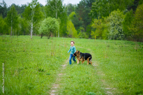 Little boy with a huge dog stands in the middle of a dirt road on a green meadow