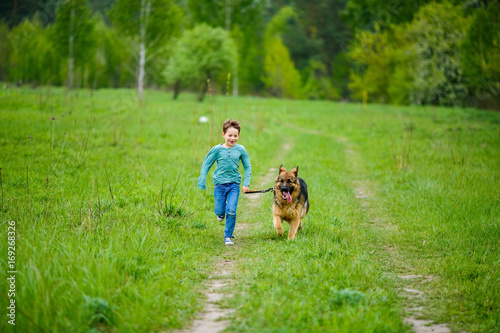 The little boy runs through the green meadow with his big dog of the breed German Shepherd