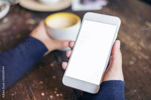 Mockup image of woman's hands holding white mobile phone with blank screen and a coffee cup on wooden table in vintage cafe